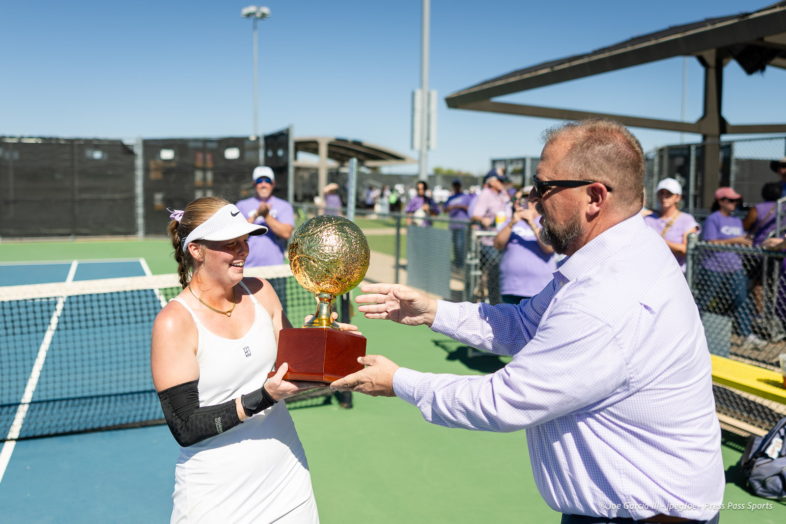 canyon-regional-tennis-105 | Press Pass Sports Tennis player in white dress receiving a gold trophy from a man in sunglasses on a sunny court, crowd cheering in background.