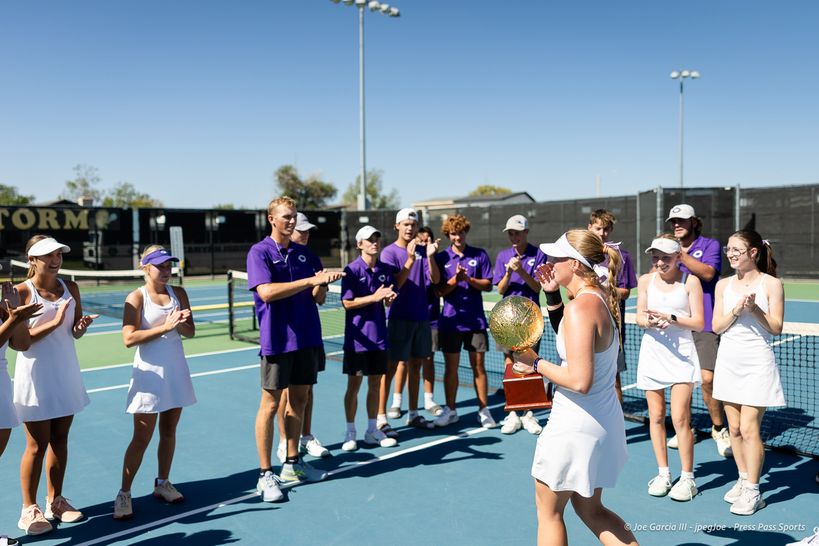 canyon-regional-tennis-106 | Press Pass Sports Tennis player in white holding a gold trophy and ball, surrounded by teammates applauding on a blue outdoor court under a clear sky