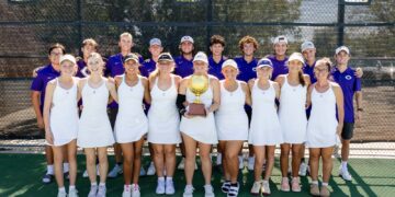 Tennis team posing on the court with a gold trophy; front row in white dresses, back row in purple uniforms.