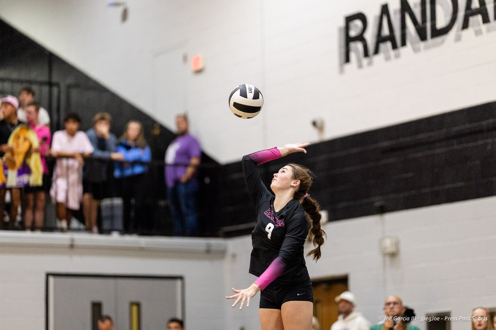 canyon-vs-west-plains-2026-3 | Press Pass Sports Young female volleyball player in black uniform with pink sleeves serves the ball in a gym, spectators watching from stands in the background