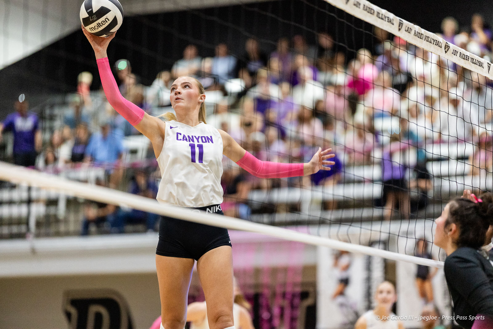 canyon-vs-west-plains-2026-6 | Press Pass Sports Volleyball player in white Canyon jersey and pink arm sleeves spikes the ball over the net with a crowd watching.