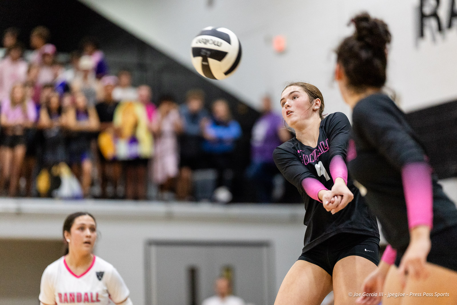 canyon-vs-west-plains-2026-7 | Press Pass Sports Volleyball player in a black uniform with pink sleeves receives or passes the ball mid-air during a match as a blurred crowd watches in the gym.
