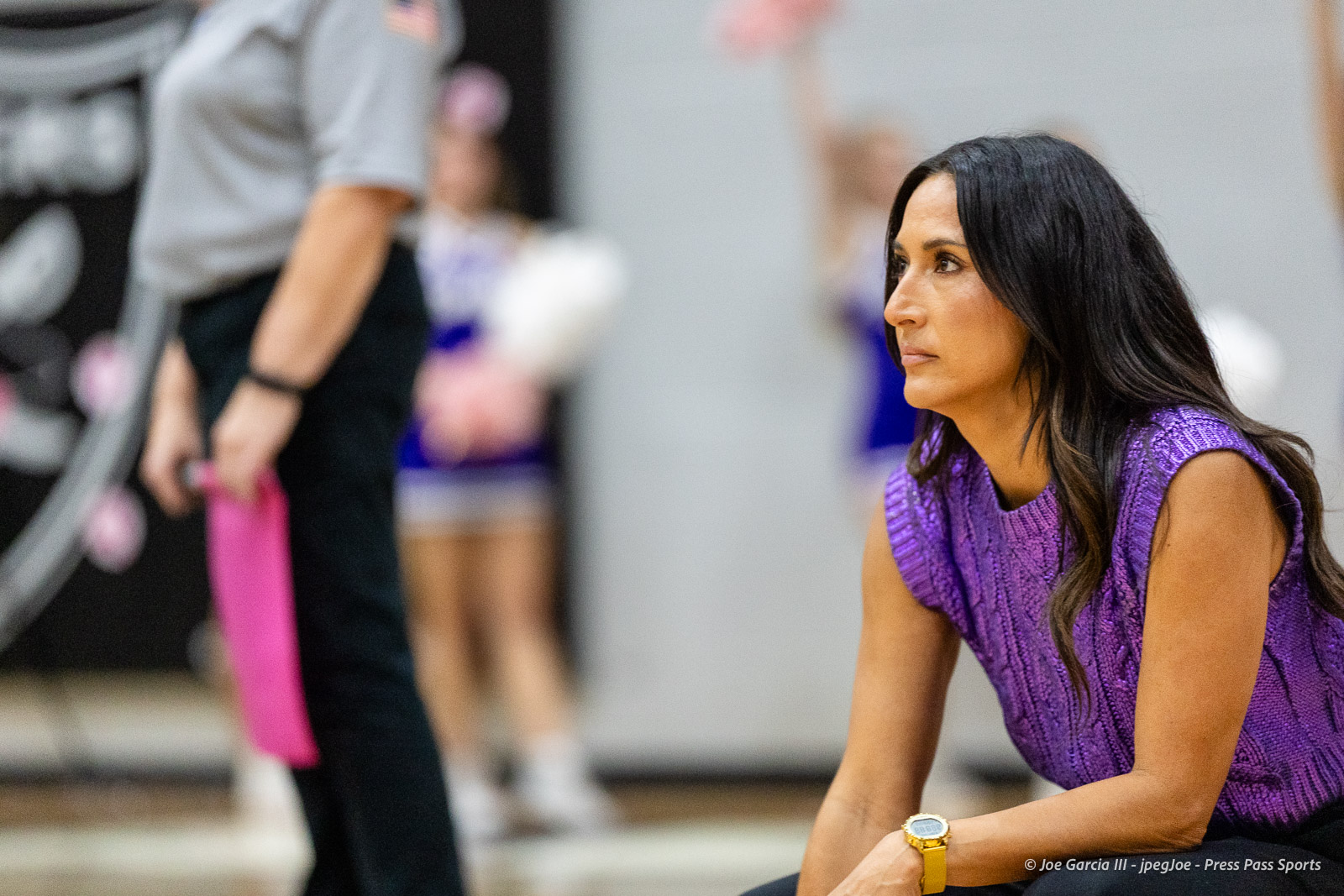canyon-vs-west-plains-2026-10 | Press Pass Sports Woman with long dark hair in a purple knit top sits on the floor, watching intently in a gym setting.