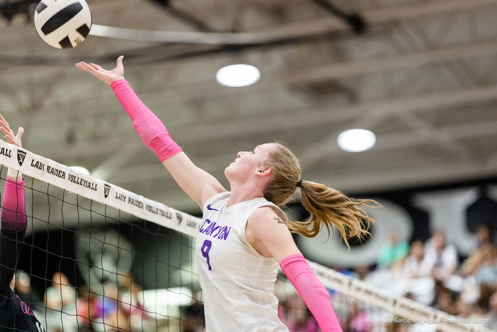 canyon-vs-west-plains-2026-13 | Press Pass Sports Female volleyball player in a white jersey (#3) with pink arm sleeves reaching to block or spike the ball at the net during a match, gym crowd in background.