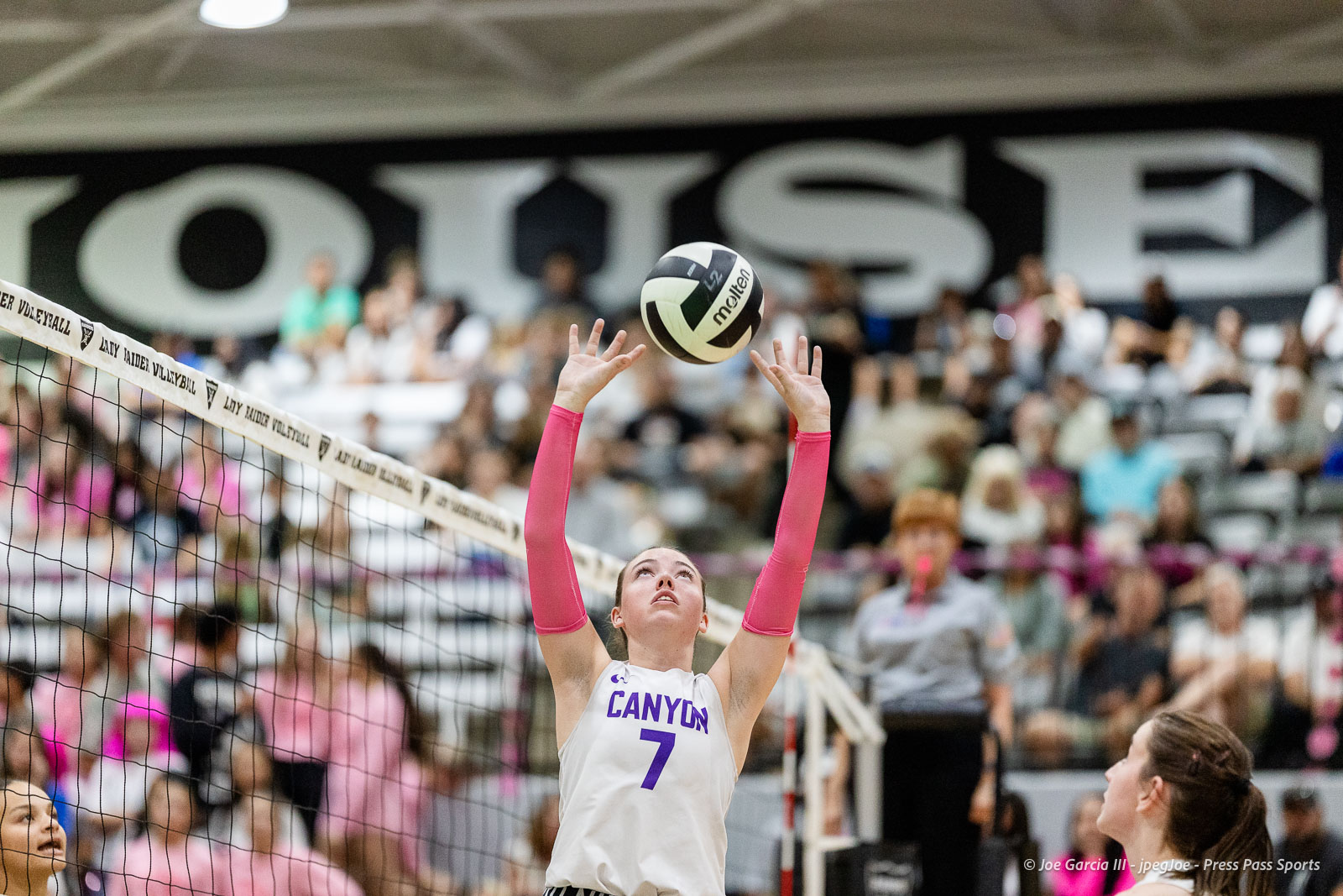 canyon-vs-west-plains-2026-14 | Press Pass Sports Volleyball player in a white Canyon jersey #7 with pink sleeves reaches up to set the ball over the net as a cheering crowd watches in the background.