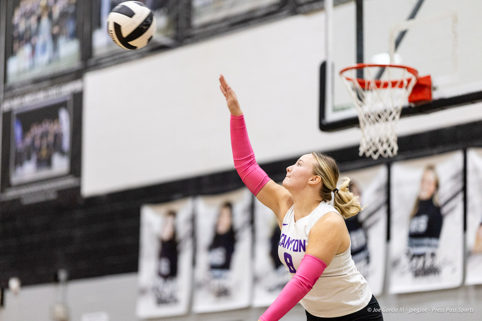 canyon-vs-west-plains-2026-15 | Press Pass Sports Female volleyball player in a white jersey serves the ball in a gym, wearing bright pink arm sleeves and ponytail.