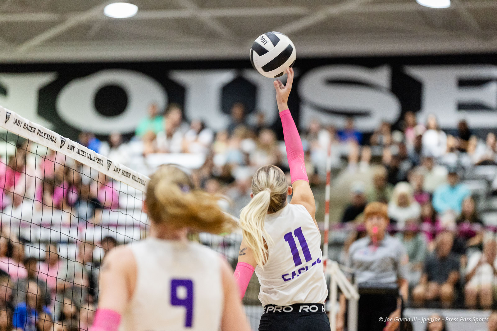 canyon-vs-west-plains-2026-16 | Press Pass Sports Female volleyball player in a white jersey with purple number 11 spikes a white and black ball over the net, pink arm sleeves, as the crowd watches from the stands behind the court.