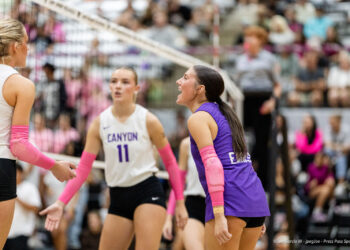 canyon-vs-west-plains-2026-18 | Press Pass Sports Female volleyball players on the court exchange a high-five near the net during a match; crowd in bleachers behind.
