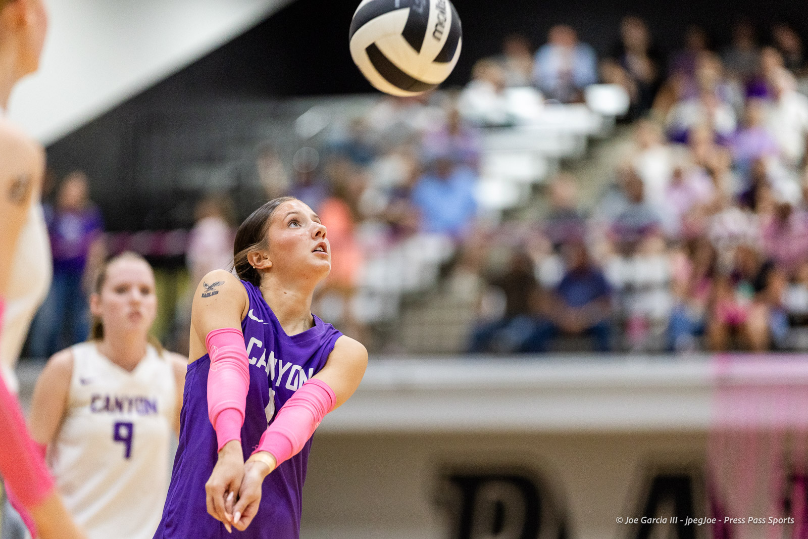 canyon-vs-west-plains-2026-19 | Press Pass Sports Female volleyball player in a purple Canyon jersey with pink sleeves reaches for the ball as it comes down, crowd in the background.