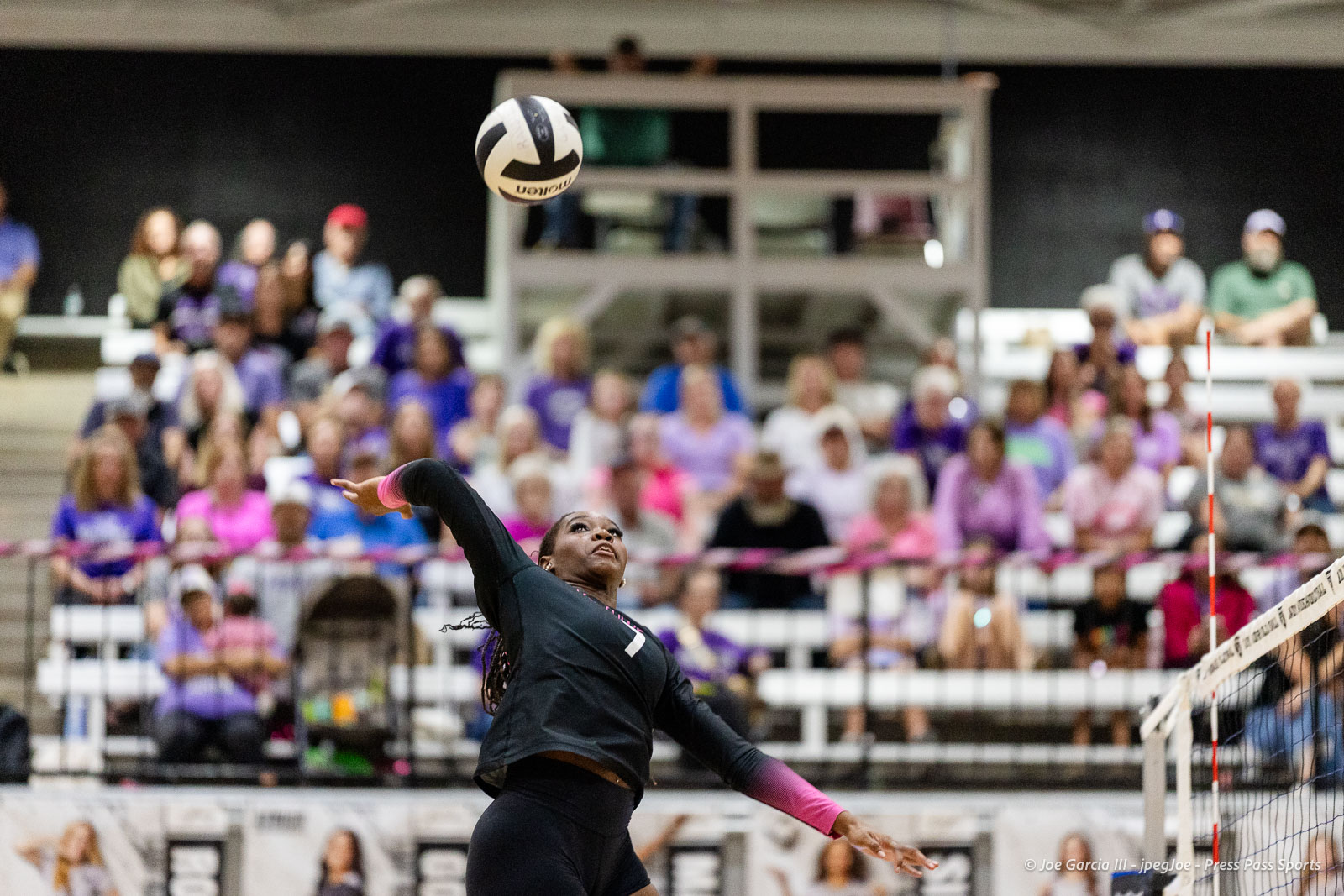 canyon-vs-west-plains-2026-22 | Press Pass Sports Female volleyball player in black with pink wristbands spikes the ball indoors, crowd watching in the stands behind her.