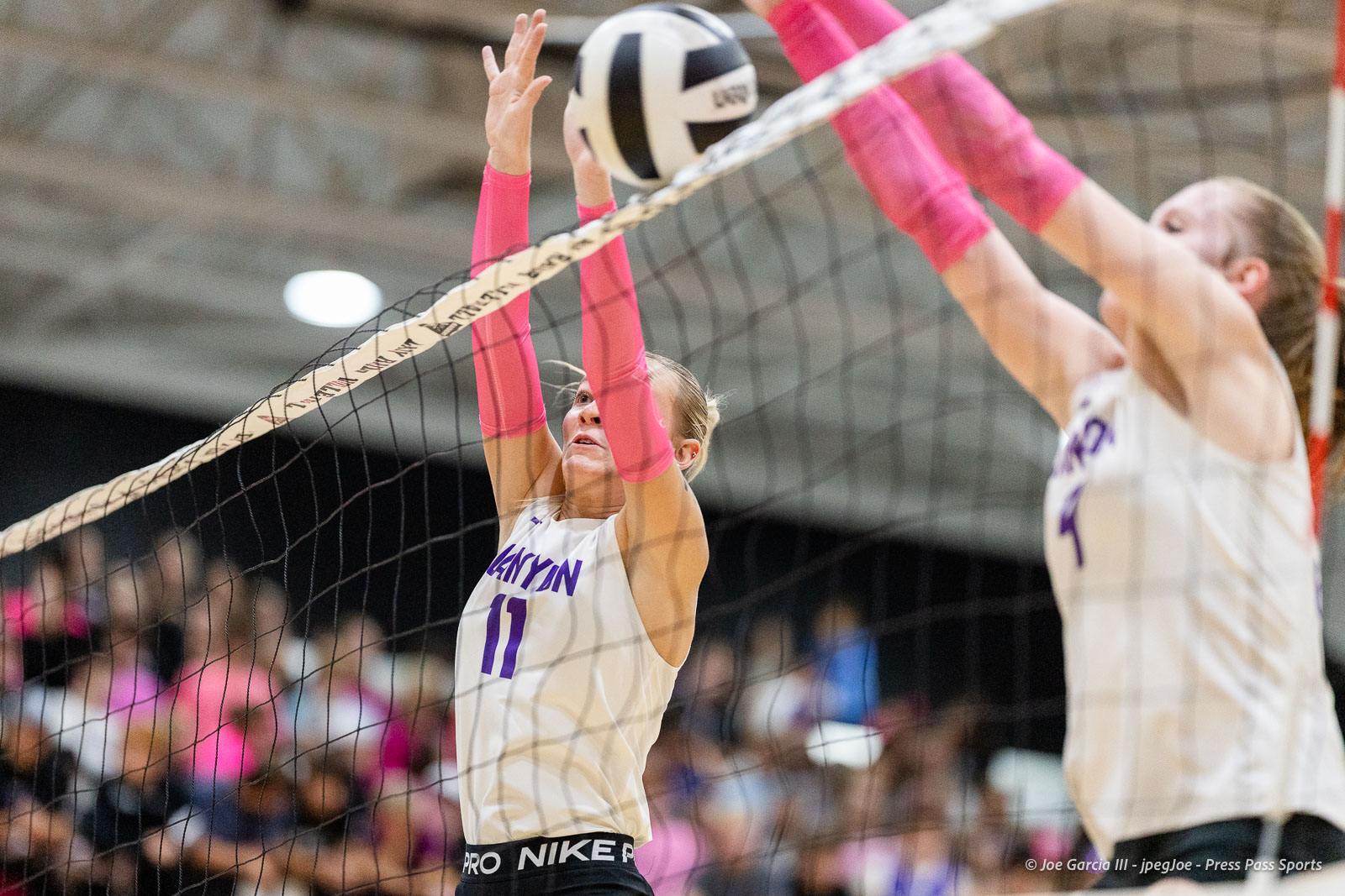 canyon-vs-west-plains-2026-23 | Press Pass Sports Female volleyball player in a white jersey with pink sleeves blocks at the net as the ball comes near.