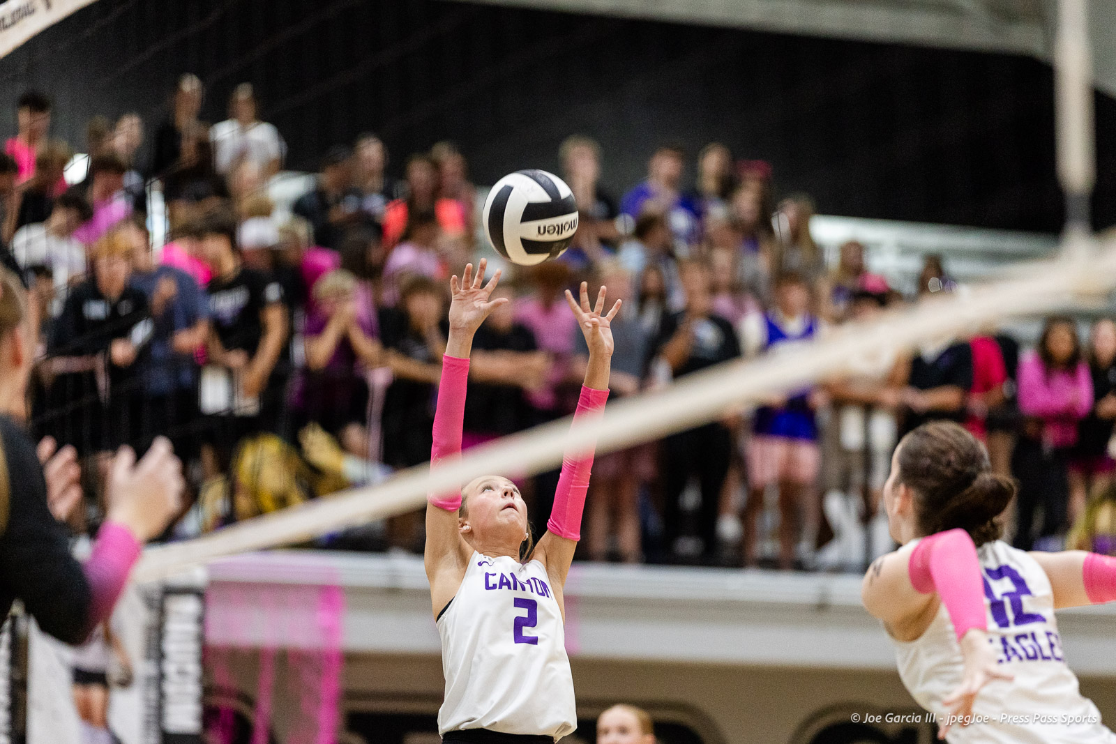 canyon-vs-west-plains-2026-24 | Press Pass Sports Female volleyball player in a white jersey number 2 with pink sleeves reaches for the ball above the net as a crowd watches.