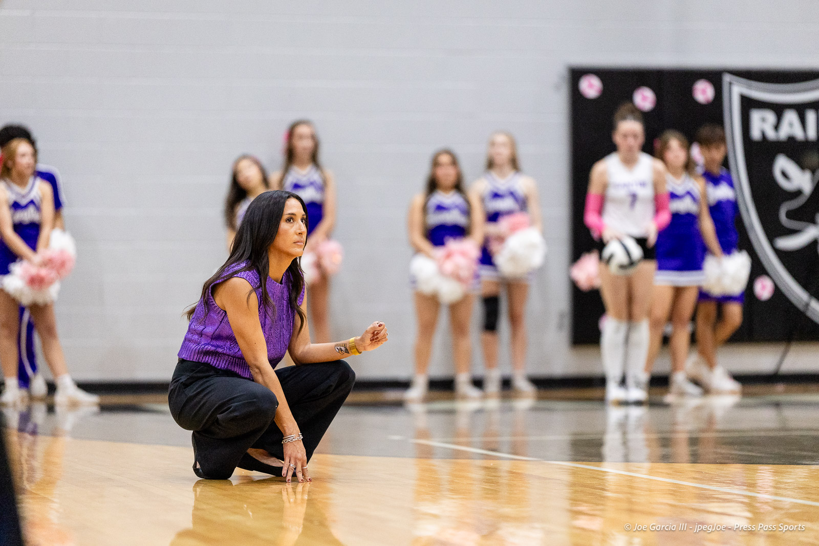 canyon-vs-west-plains-2026-26 | Press Pass Sports Coach kneeling on a basketball court in a purple sleeveless top, with blurred cheerleaders in purple and white uniforms in the background.