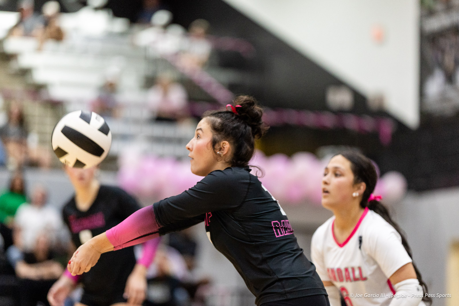 canyon-vs-west-plains-2026-28 | Press Pass Sports Athlete in a black uniform with pink sleeves extends her arms to hit a volleyball, with a teammate in white watching in the background during an indoor match.