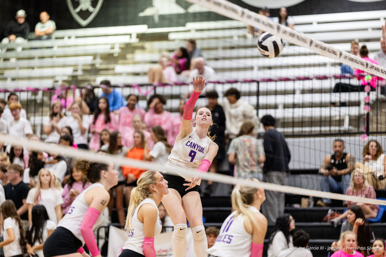 canyon-vs-west-plains-2026-29 | Press Pass Sports Female volleyball player (CANYON #9) jumps to strike the ball at the net as fans watch from the bleachers, pink sleeves on both arms nearby.