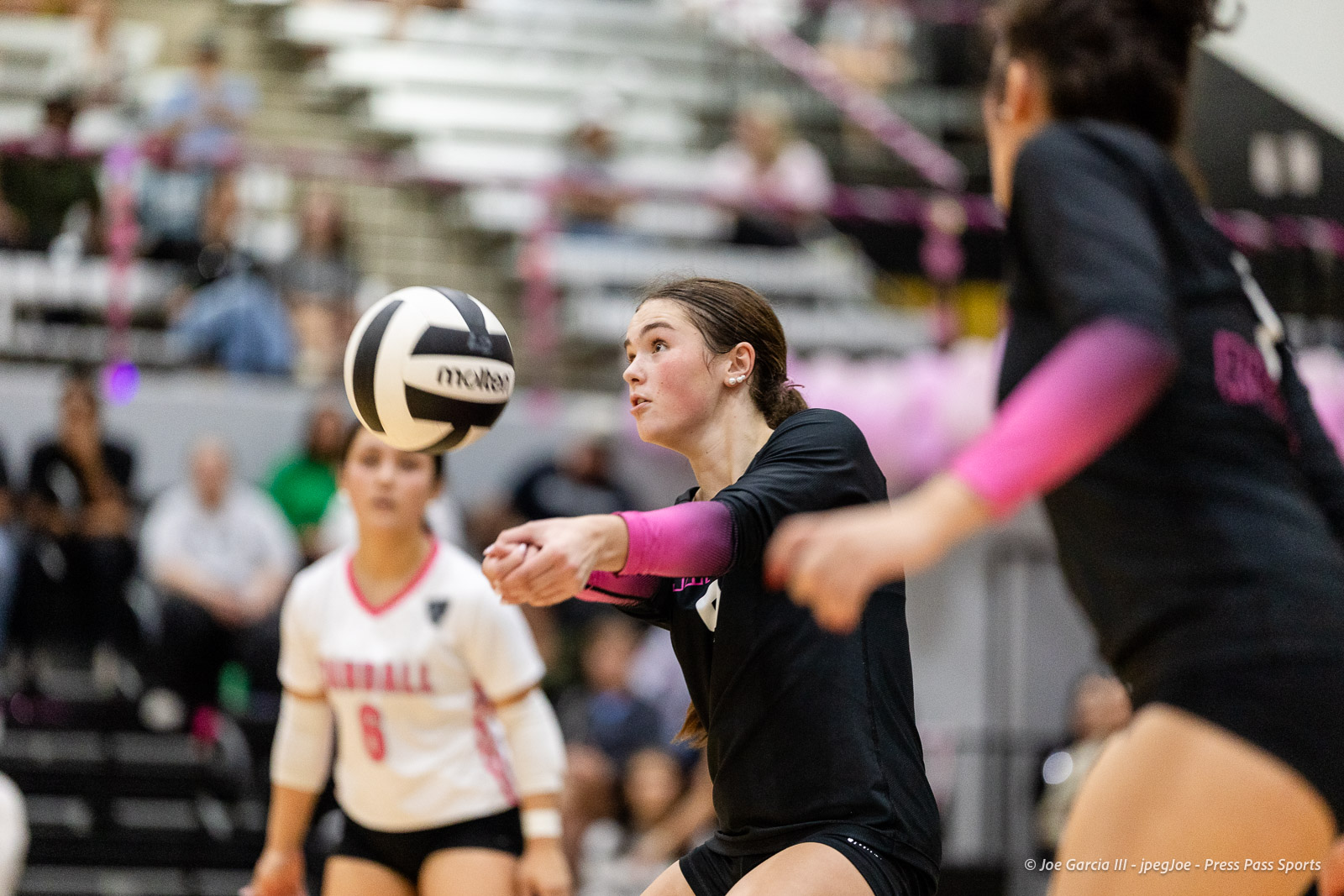 canyon-vs-west-plains-2026-30 | Press Pass Sports Female volleyball player in black uniform and pink sleeves dives to receive the ball during a match in a gymnasium with blurred spectators in the background.