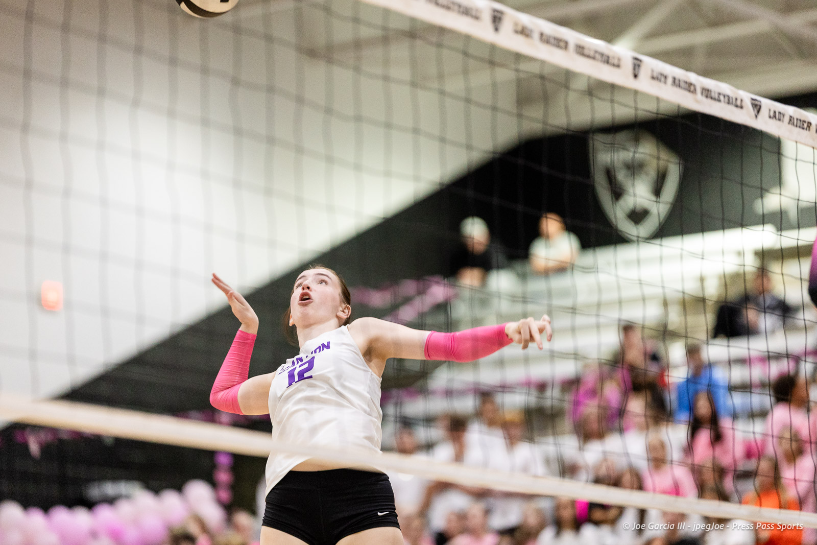 canyon-vs-west-plains-2026-31 | Press Pass Sports Female volleyball player in a white jersey and pink arm sleeves spikes the ball over the net with spectators in the background.