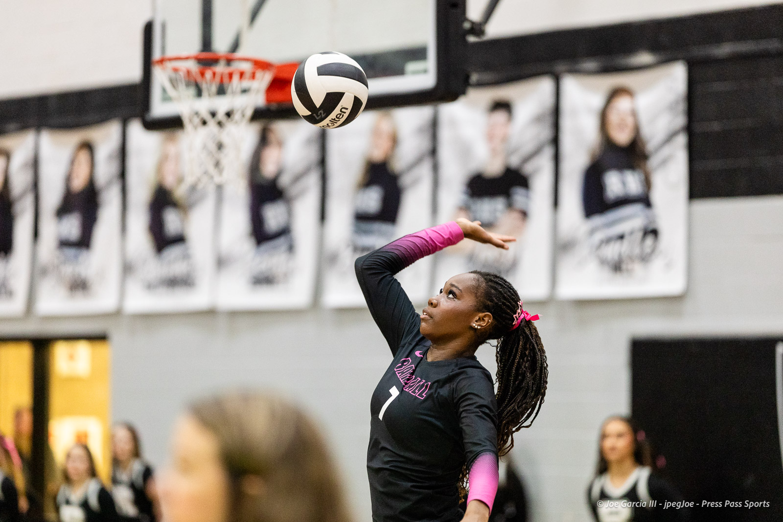 canyon-vs-west-plains-2026-33 | Press Pass Sports Female volleyball player in a black jersey with pink sleeves serves the ball in a gym.