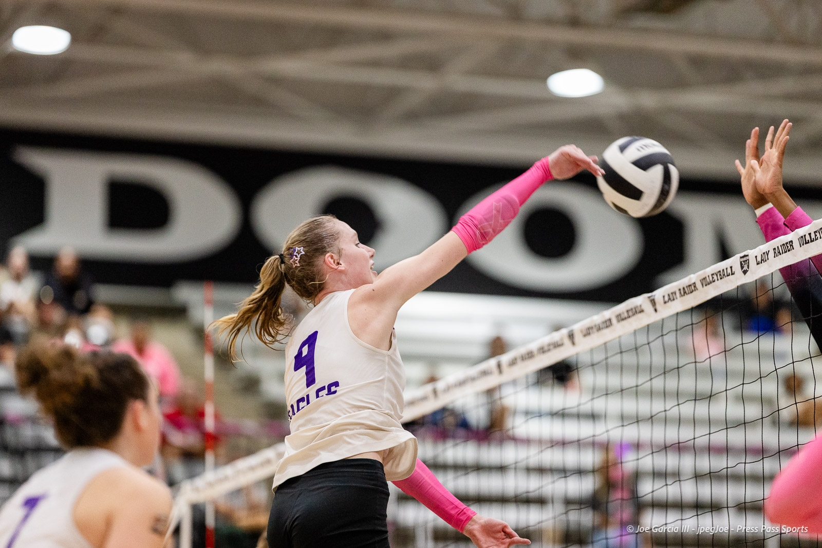 canyon-vs-west-plains-2026-37 | Press Pass Sports Female volleyball player in a white jersey with pink sleeves spikes the ball at the net during a match.
