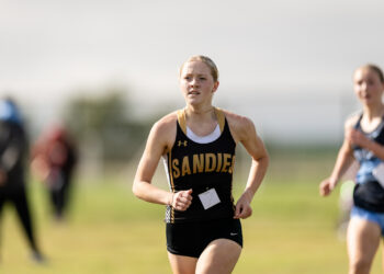 5a-2026-district-cross-country-xc-jpegJoe-pps-33 | Press Pass Sports Female runner in a black and gold singlet races on a sunny field, bib attached to front.