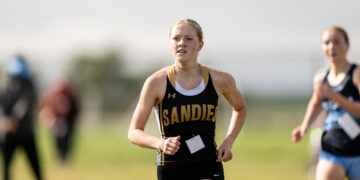 Female runner in a black and gold singlet races on a sunny field, bib attached to front.
