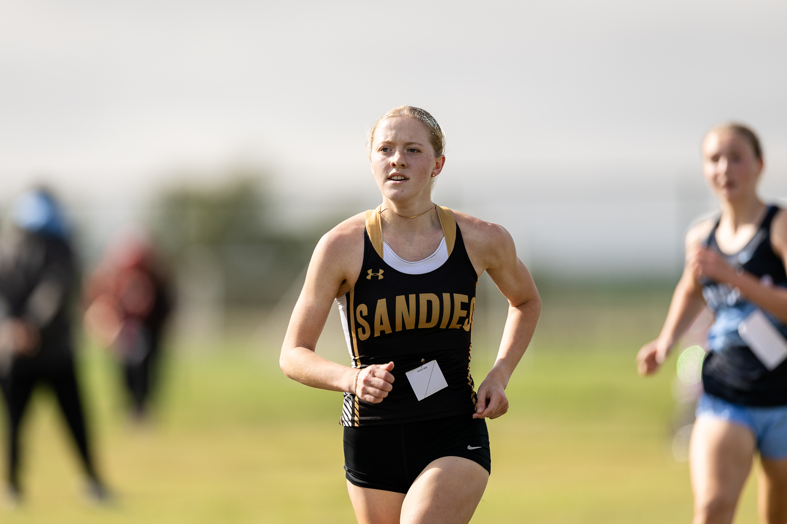 5a-2026-district-cross-country-xc-jpegJoe-pps-33 | Press Pass Sports Female runner in a black and gold singlet races on a sunny field, bib attached to front.