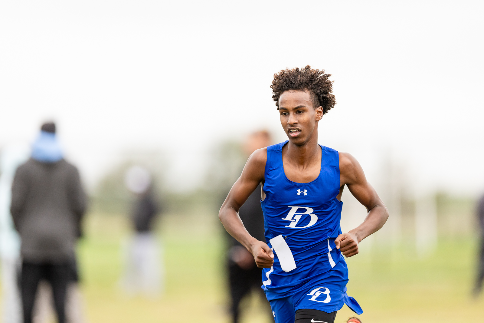 5a-2026-district-cross-country-xc-jpegJoe-pps-16 | Press Pass Sports Male sprinter in a blue BD uniform sprints on an outdoor field, holding a white race bib tag in his hand under a cloudy sky.