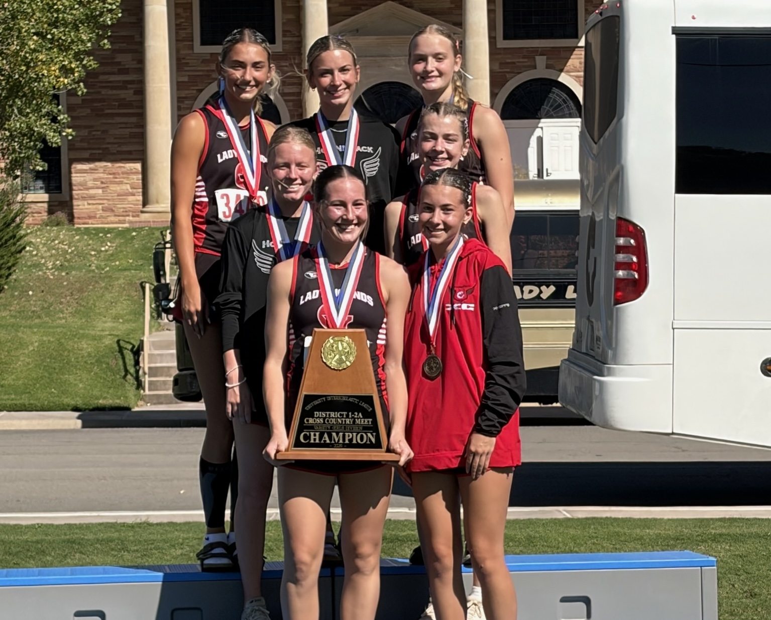 IMG_8835 | Press Pass Sports Group of young female runners posing on a podium with medals and a large champion trophy front and center.