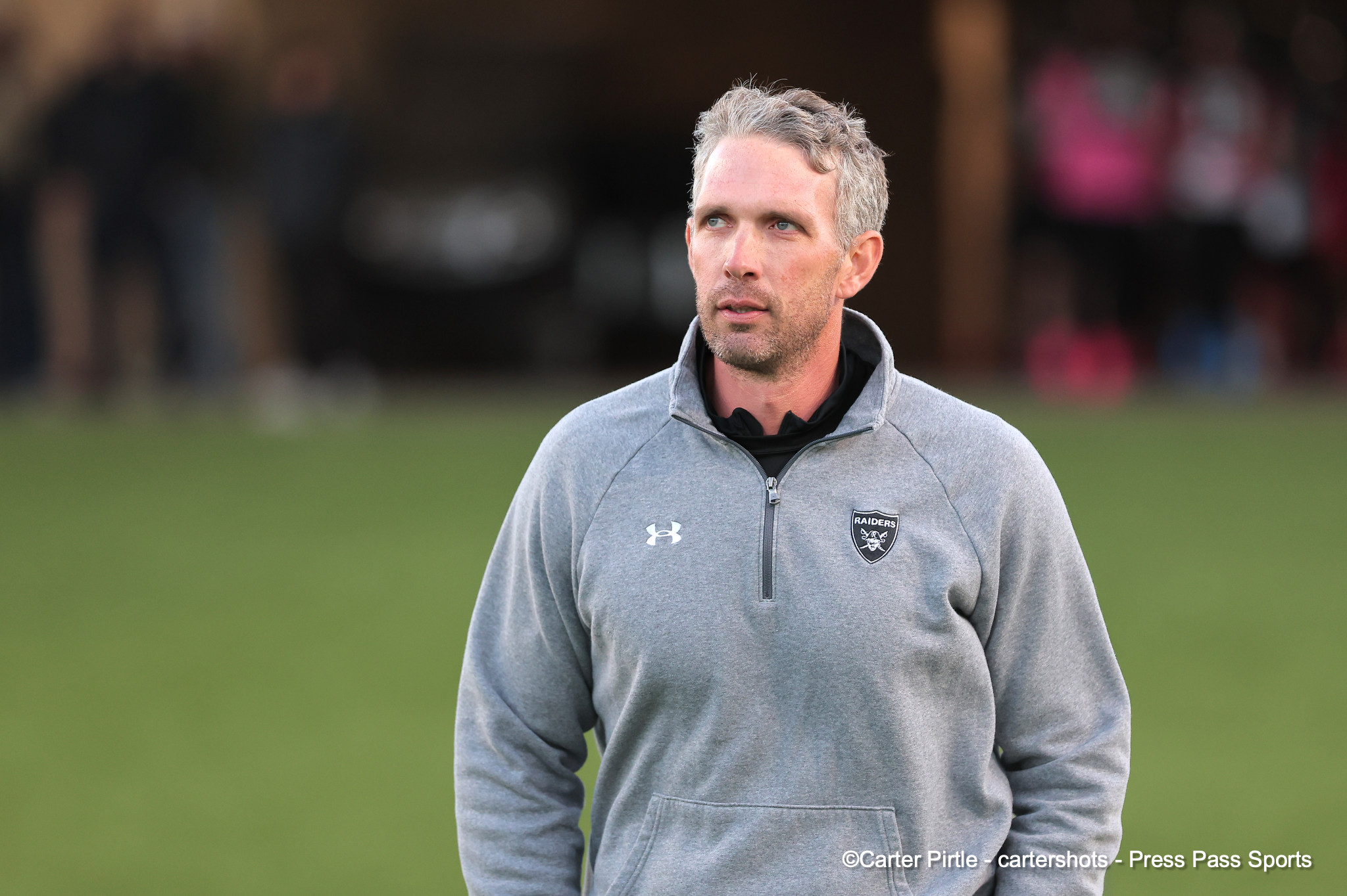 Man standing outdoors on a grassy field, wearing a gray Raiders zip-up jacket and looking to the right, with a blurred background.