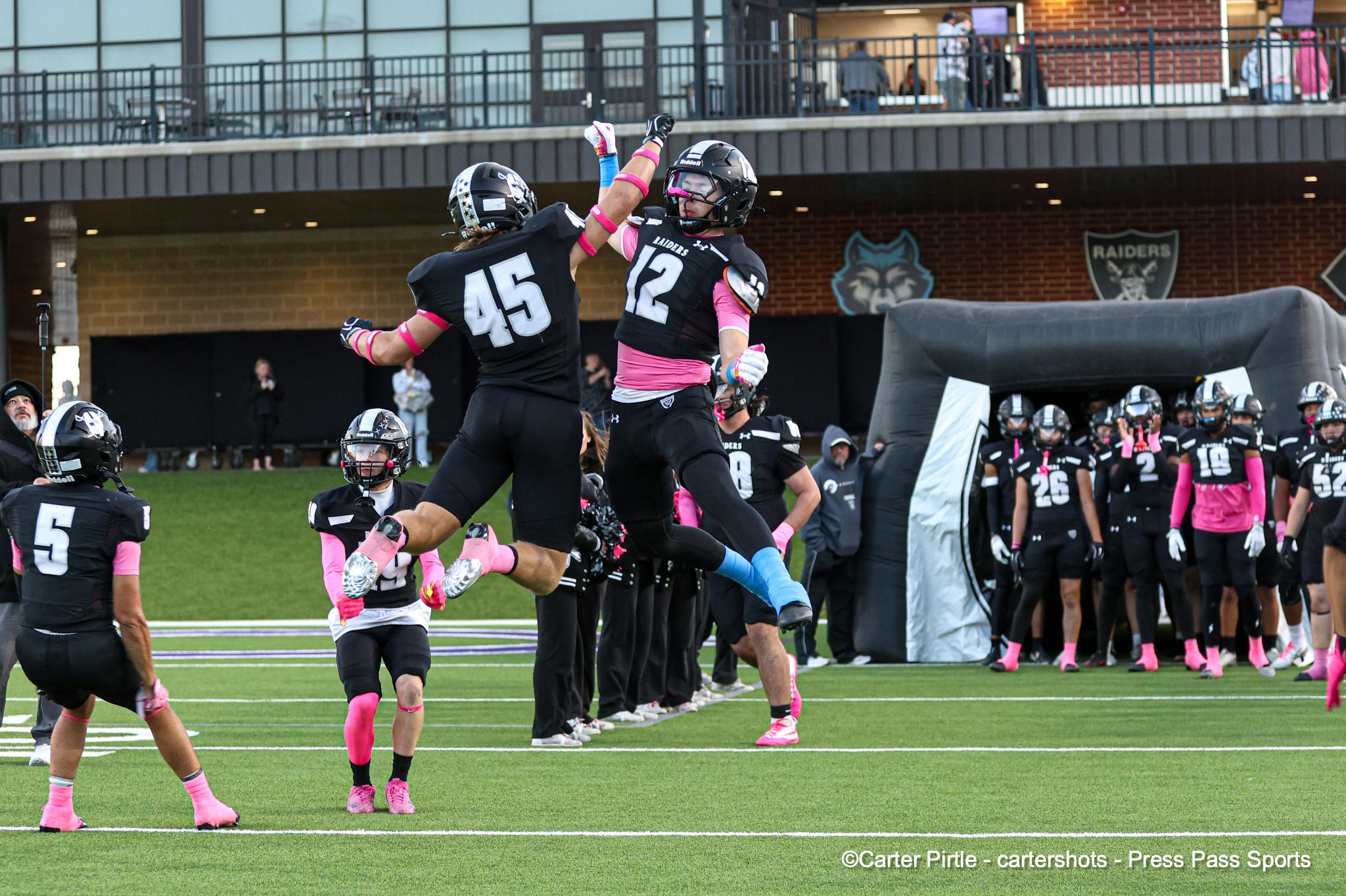 Two football players in black and pink jump midair to high-five on the field during a game.
