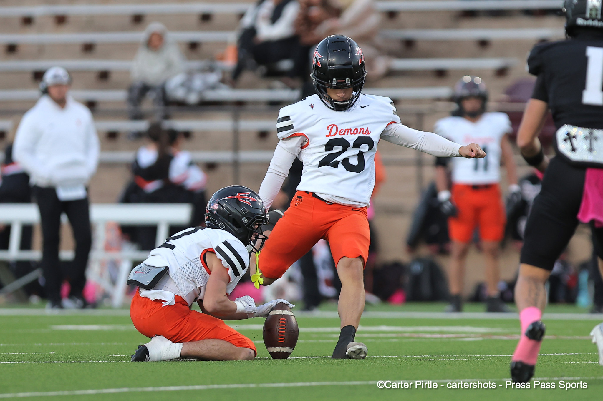 Football player in a white Demons jersey (#23) leaps over a fallen opponent during a play on a green turf field.