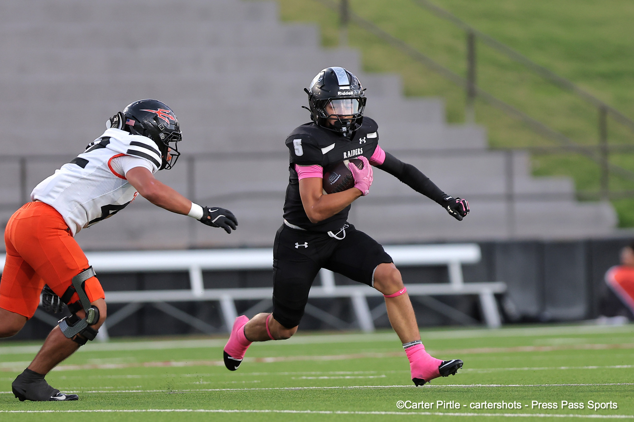 Black Raiders player with the ball runs forward while a white-and-orange defender closes in on a green football field.