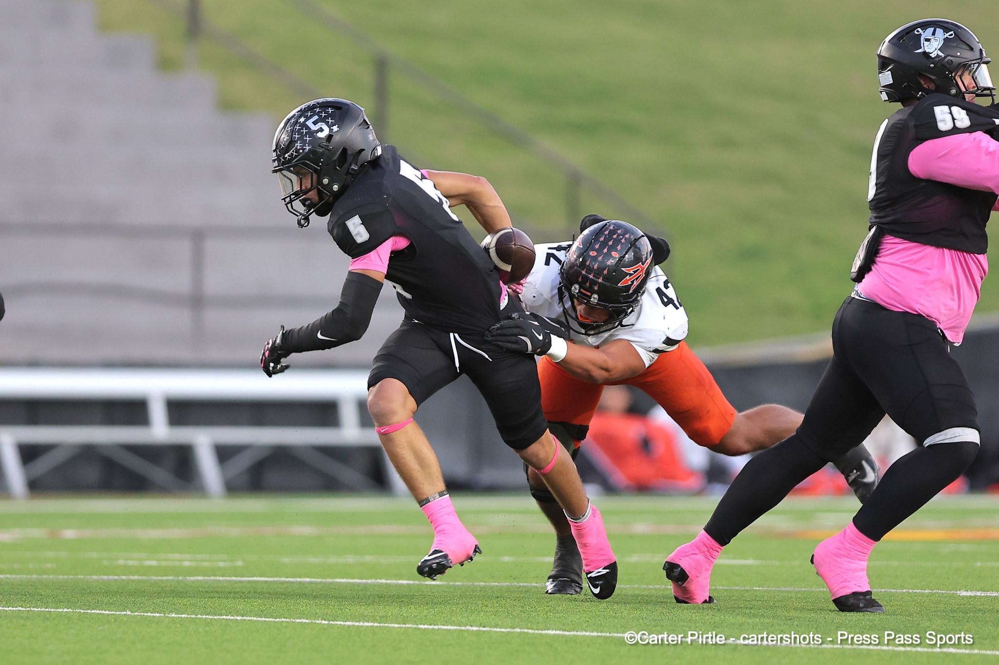 Football players in black and pink uniforms collide during a game on a green field; a tackler grabs a ball carrier.