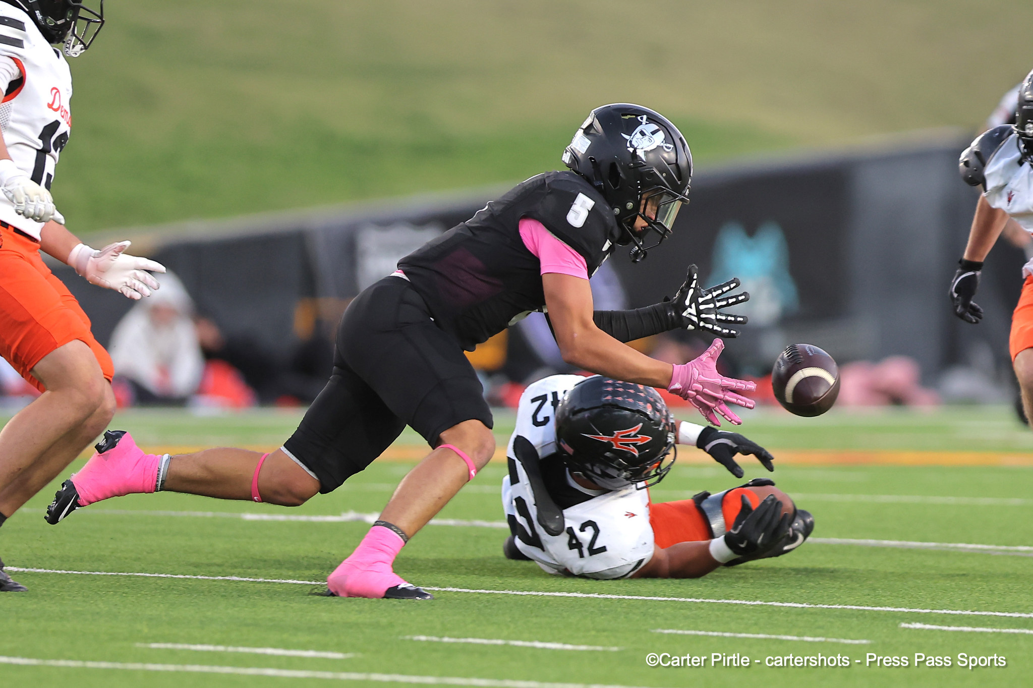 Player in black uniform (#5) reaches for a loose football as opponents in white/orange close by on the turf field. Pink gloves stand out.