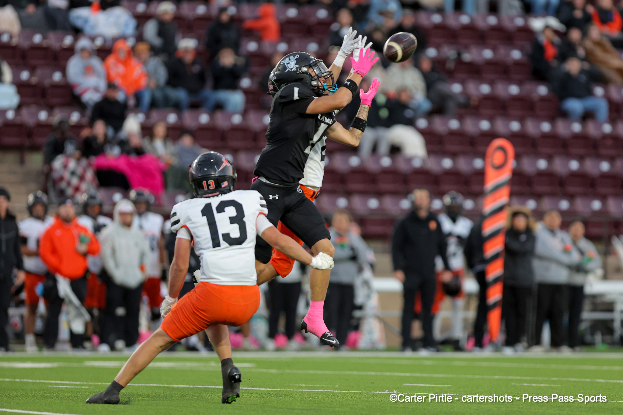 Two football players jump for a spiraling ball on a green field with a cheering crowd in the stands behind them.