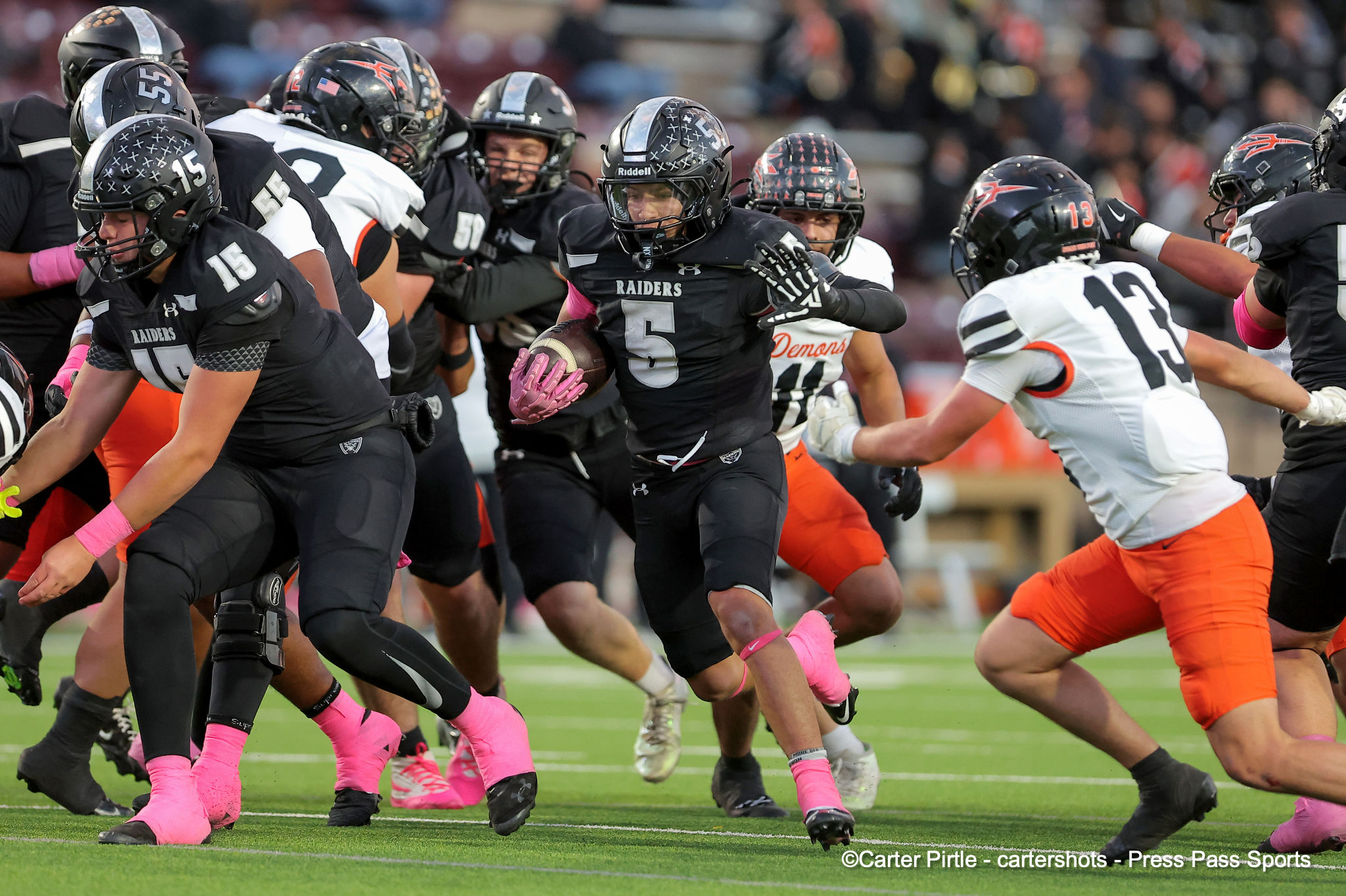 Raiders running back in black jersey #5 cradles the ball as teammates and defenders clash around him on the field.