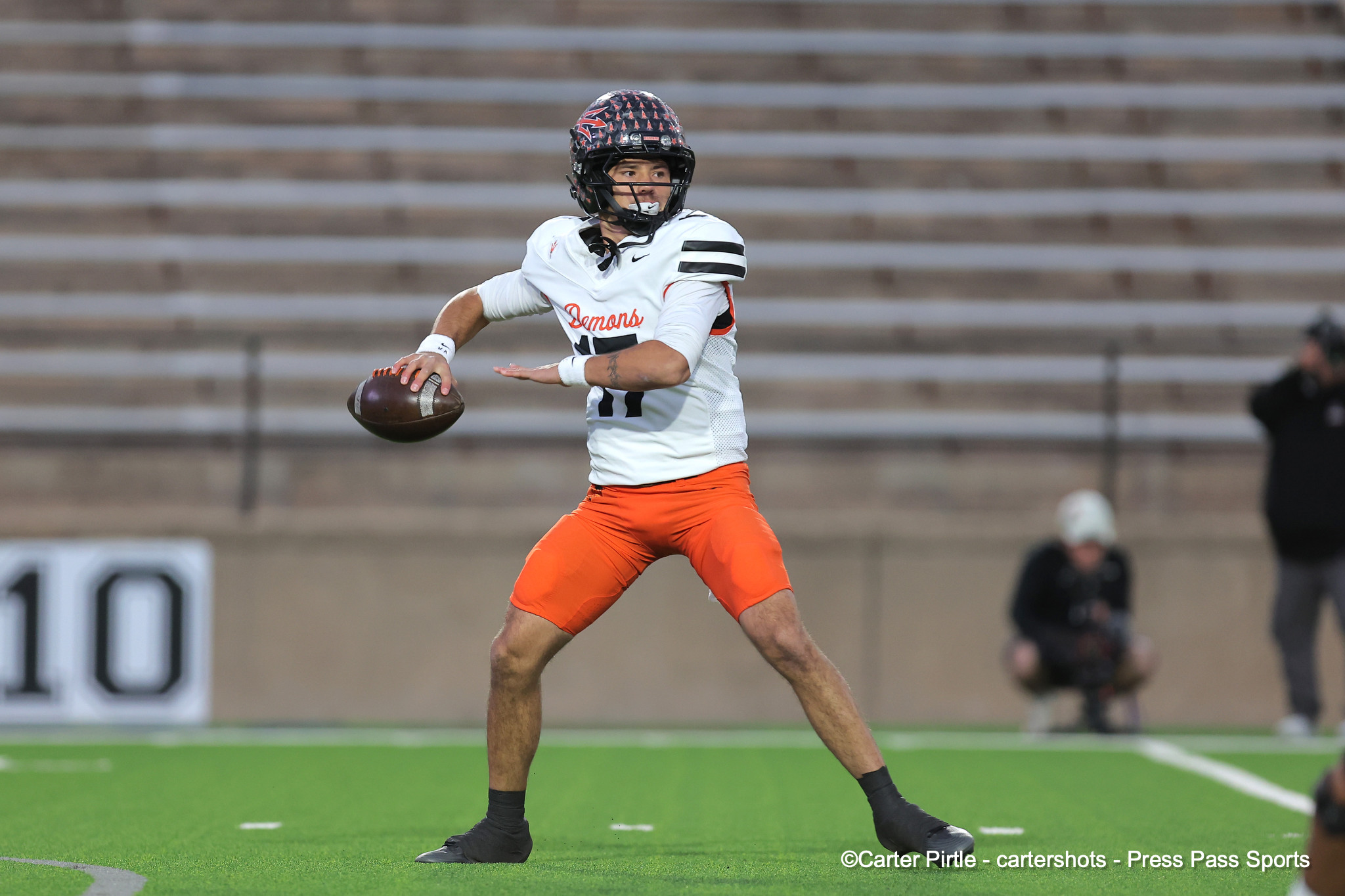 Quarterback in a white Demons jersey and orange pants throwing a football on a green turf field