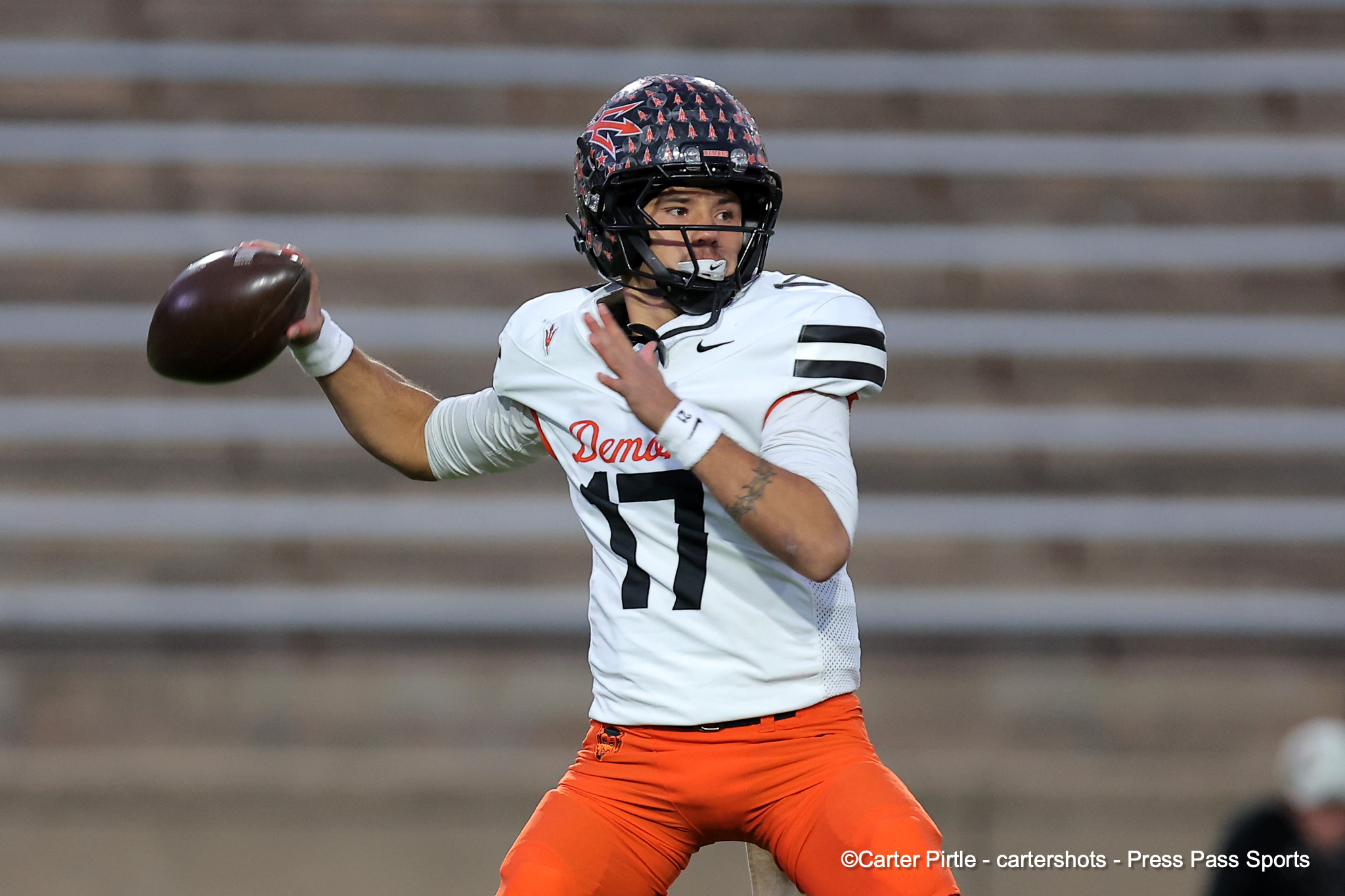 Quarterback in a white jersey with orange pants throws a football during a game, wearing a patterned helmet and black faceguard.