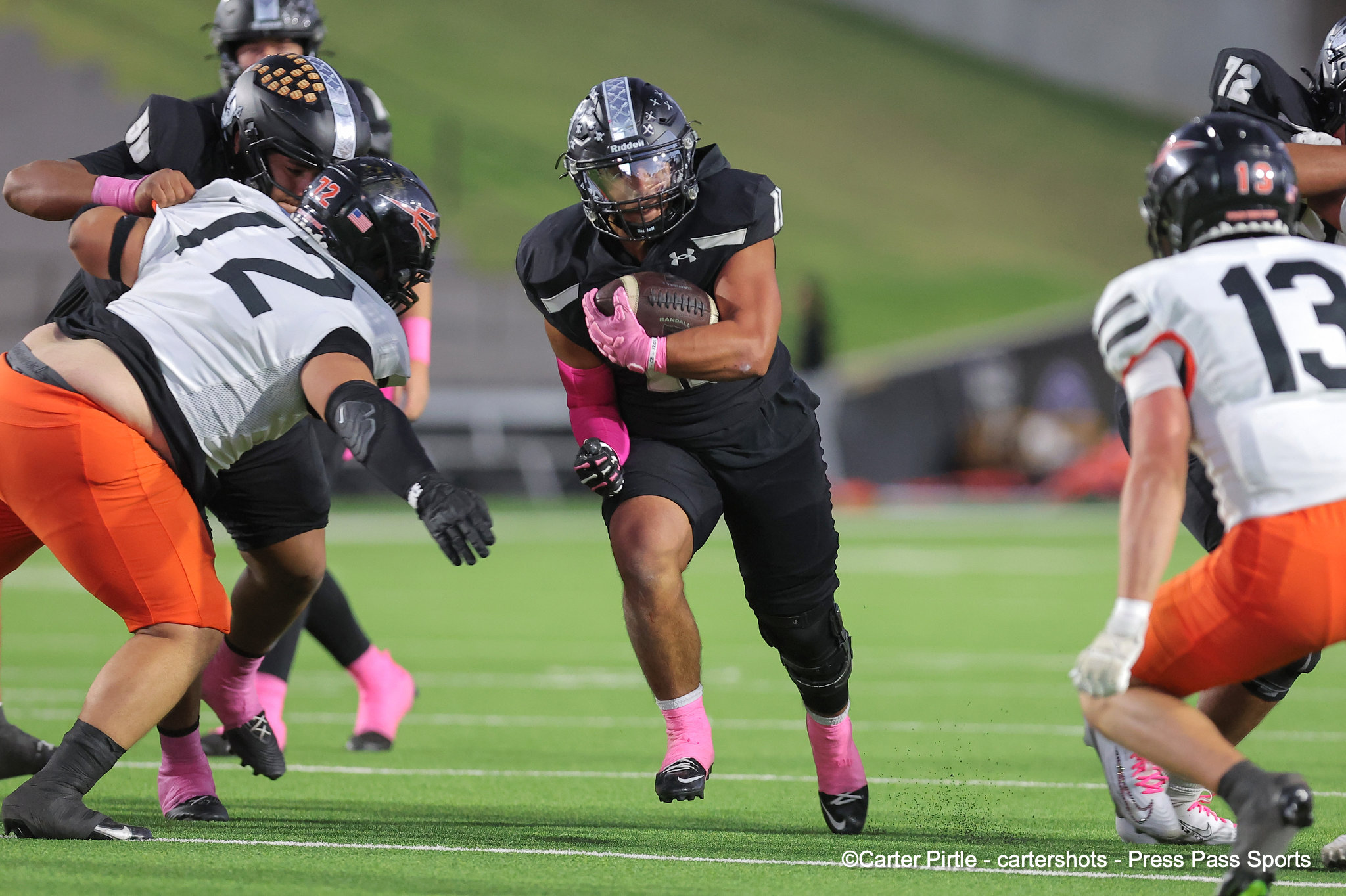 Black-uniform football player with pink gloves runs with the ball as defenders close in on a green turf field.