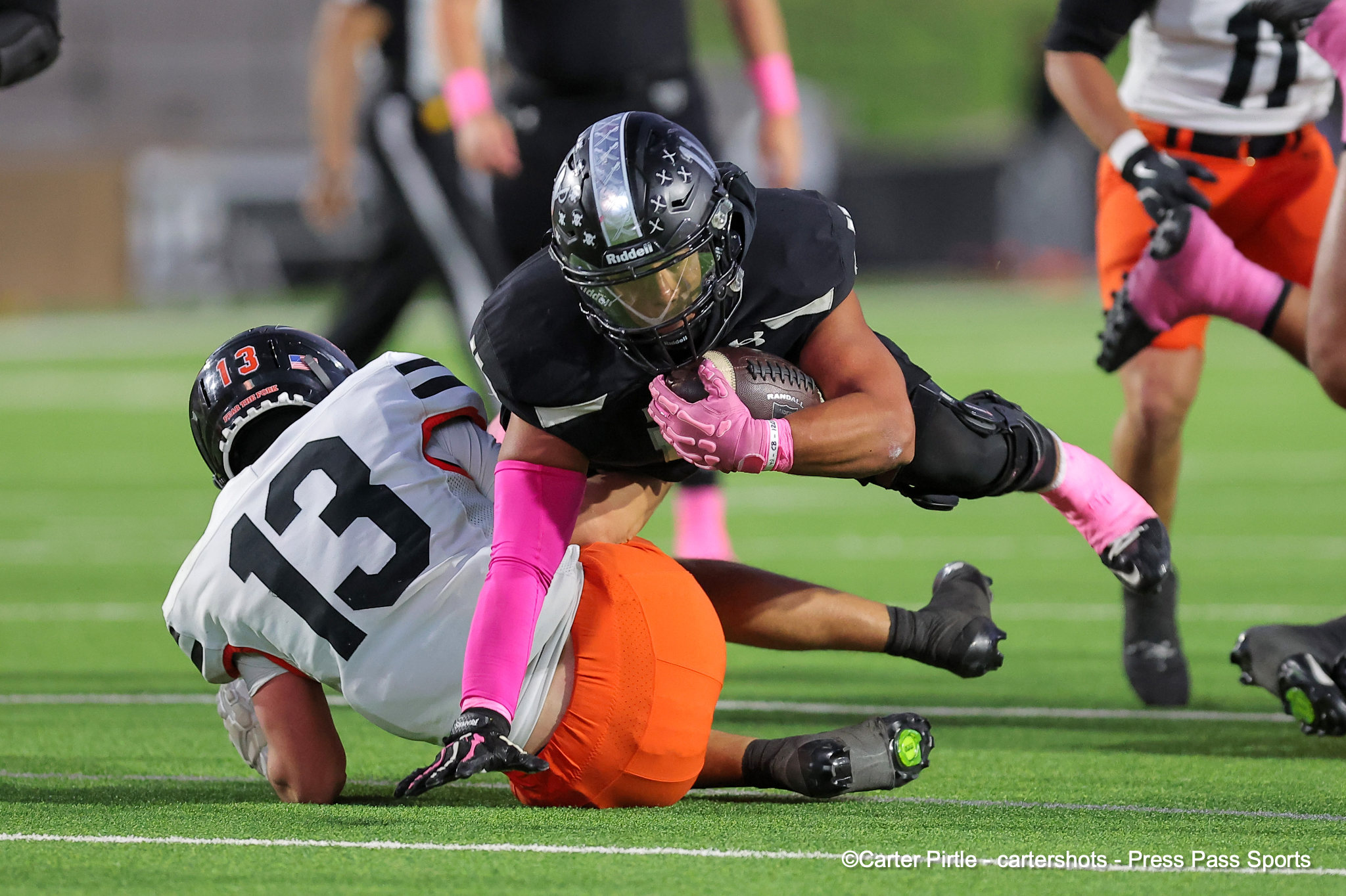 Two football players collide during a tackle on a turf field; one in black, the other in white with number 13 on the helmet, pink gloves visible.