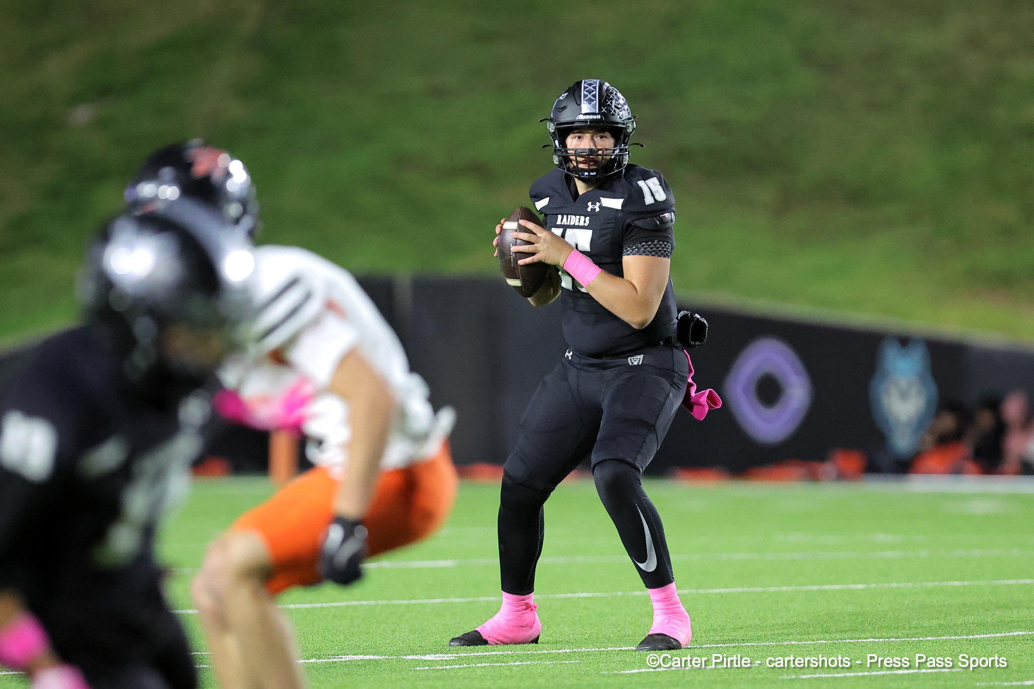 Quarterback in a black uniform (number 15) holding a football and preparing to throw on a green field, pink socks visible.