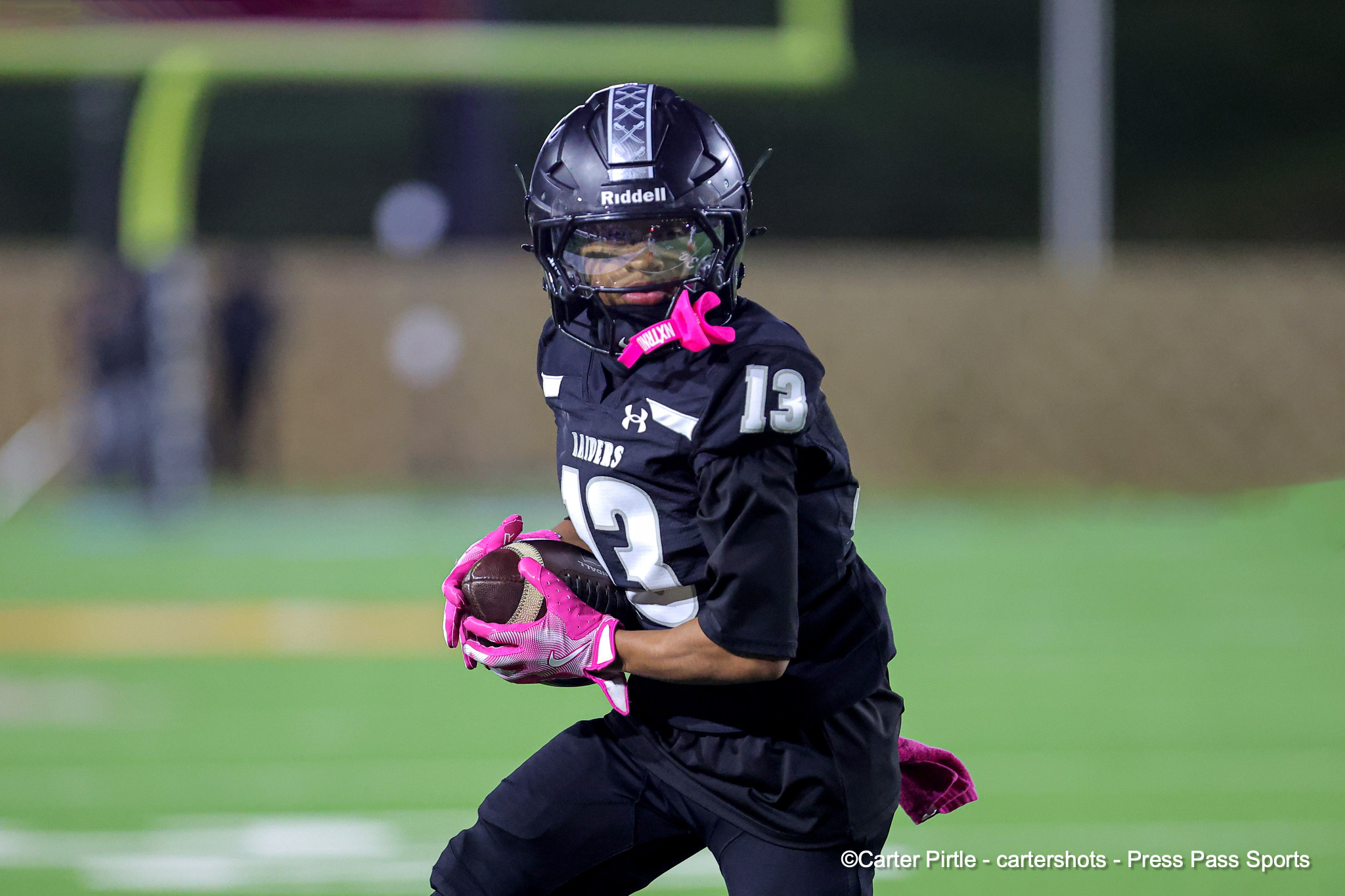 Lacrosse player in a black helmet and jersey with number 13, pink gloves, holding a brown lacrosse ball on a green field.