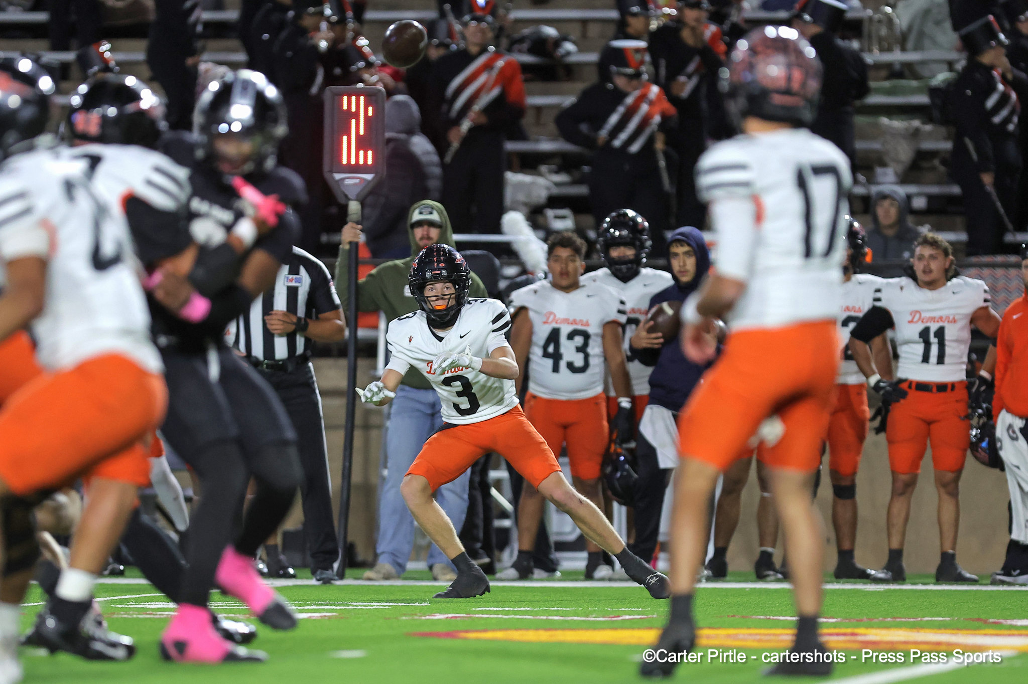 Football player wearing white jersey #3 runs with the ball on a green turf as teammates and opponents close in, stands and crowd in background.
