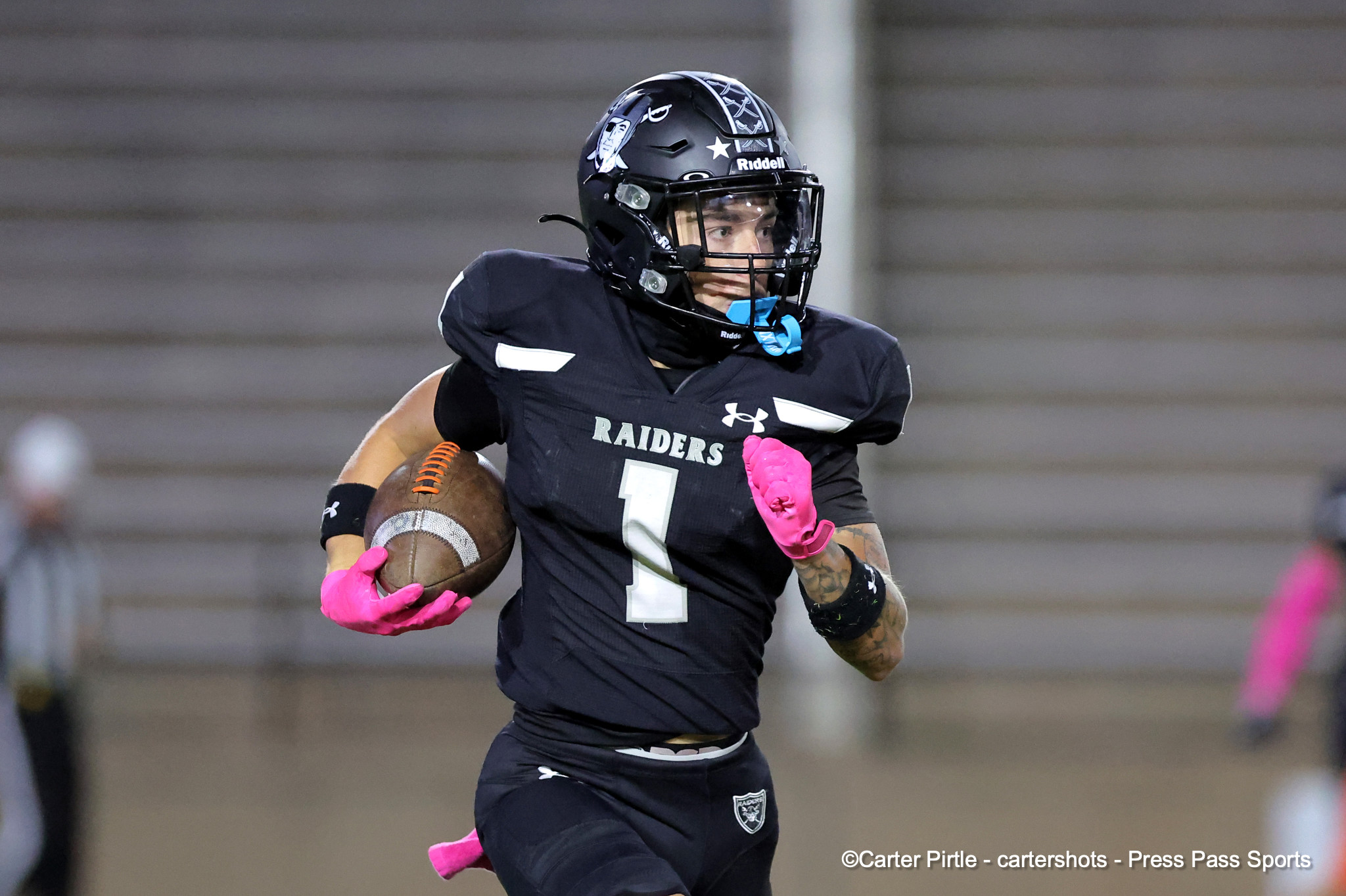 Football player in a black Raiders uniform runs with the ball, wearing a black helmet and pink gloves.