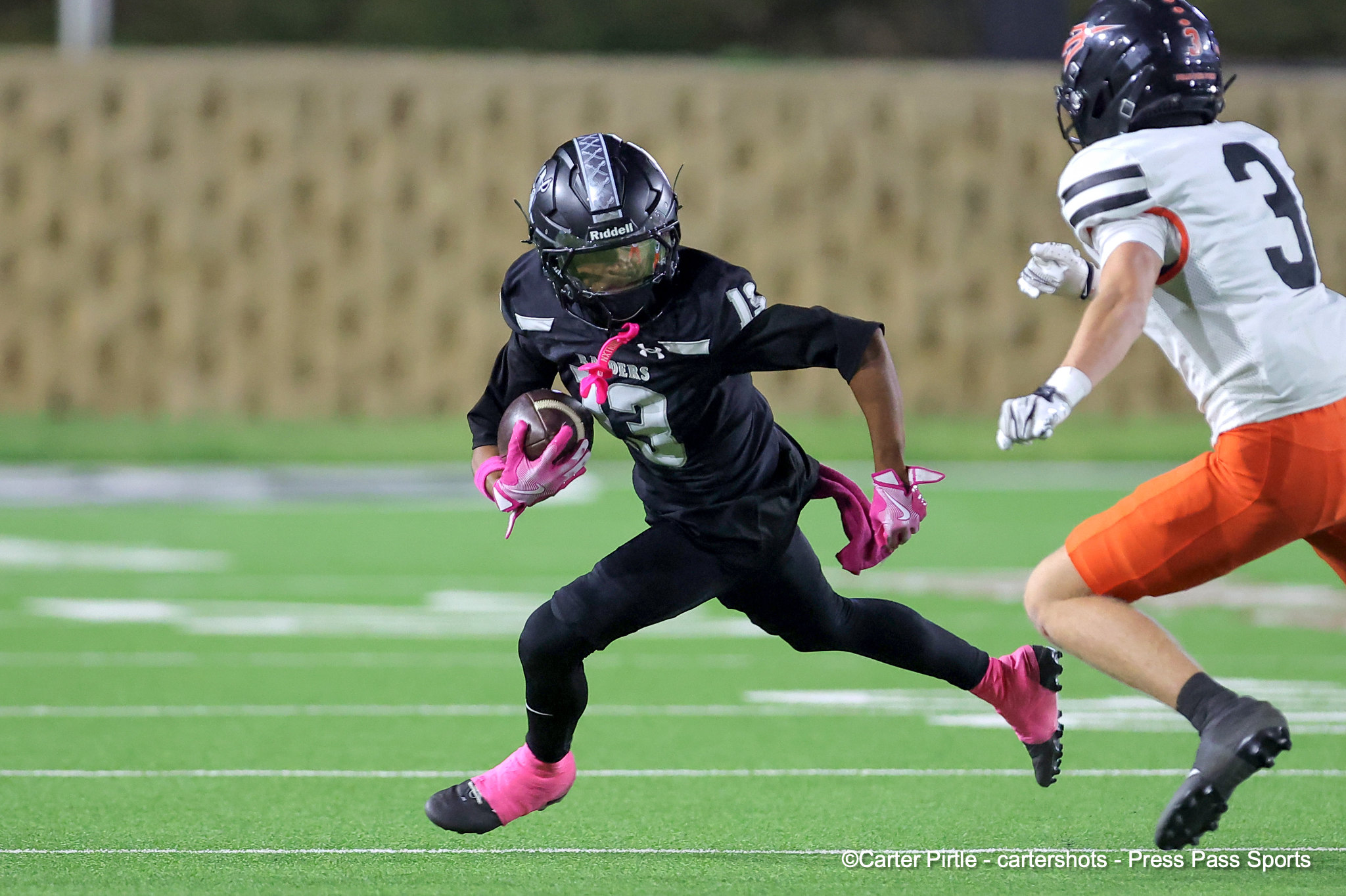 Boy in a black jersey running with the football, pink gloves, chased by a defender in a white jersey on a green turf field.