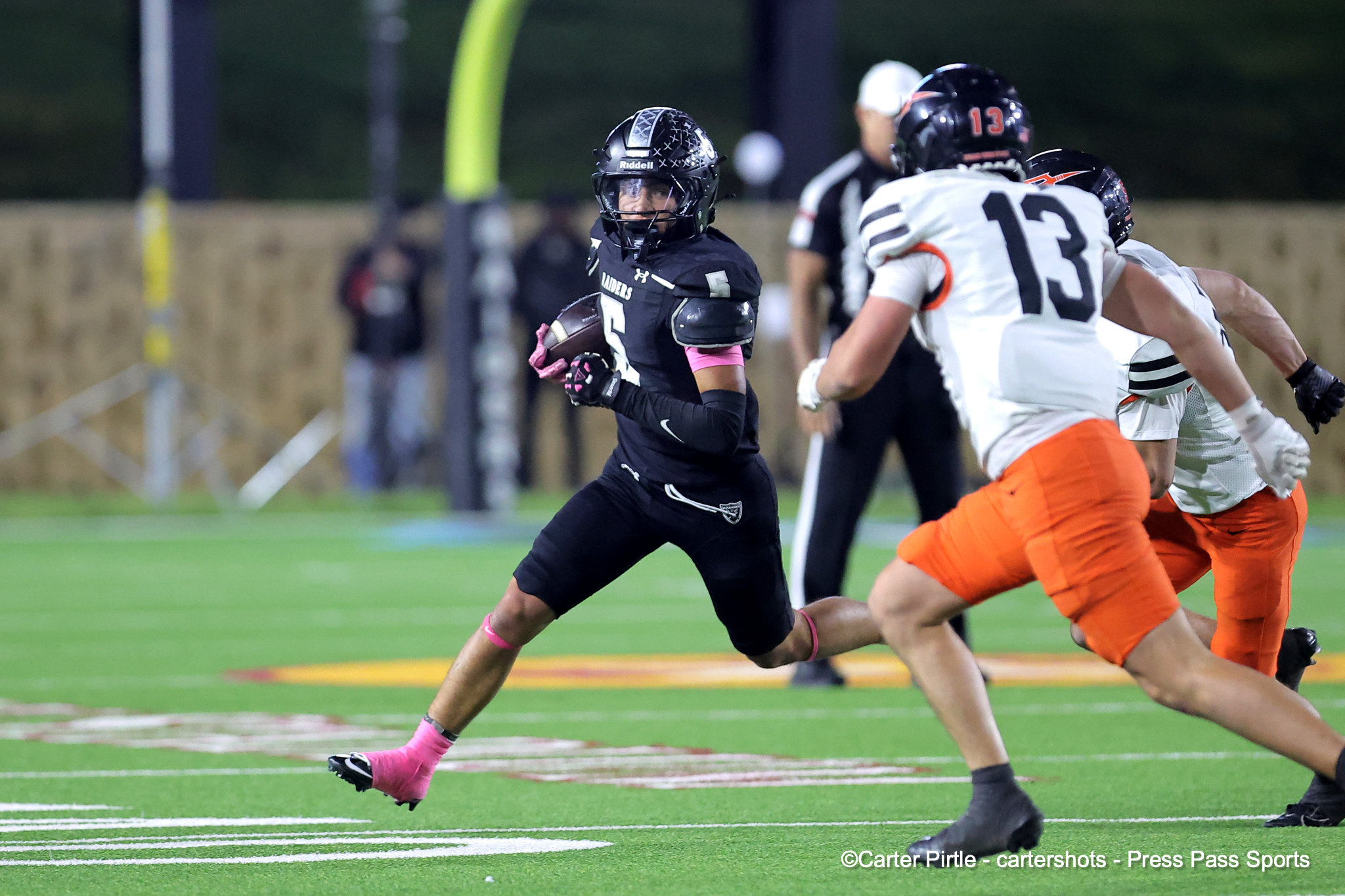 Football player in a black uniform (number 5) carrying the ball races forward as white-and-orange defenders chase on a green turf field.