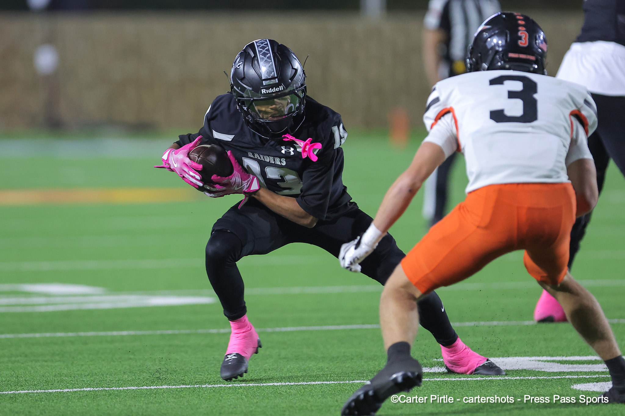Football player in a black Raiders uniform runs with the ball, pursued by a defender in white and orange on a green field.