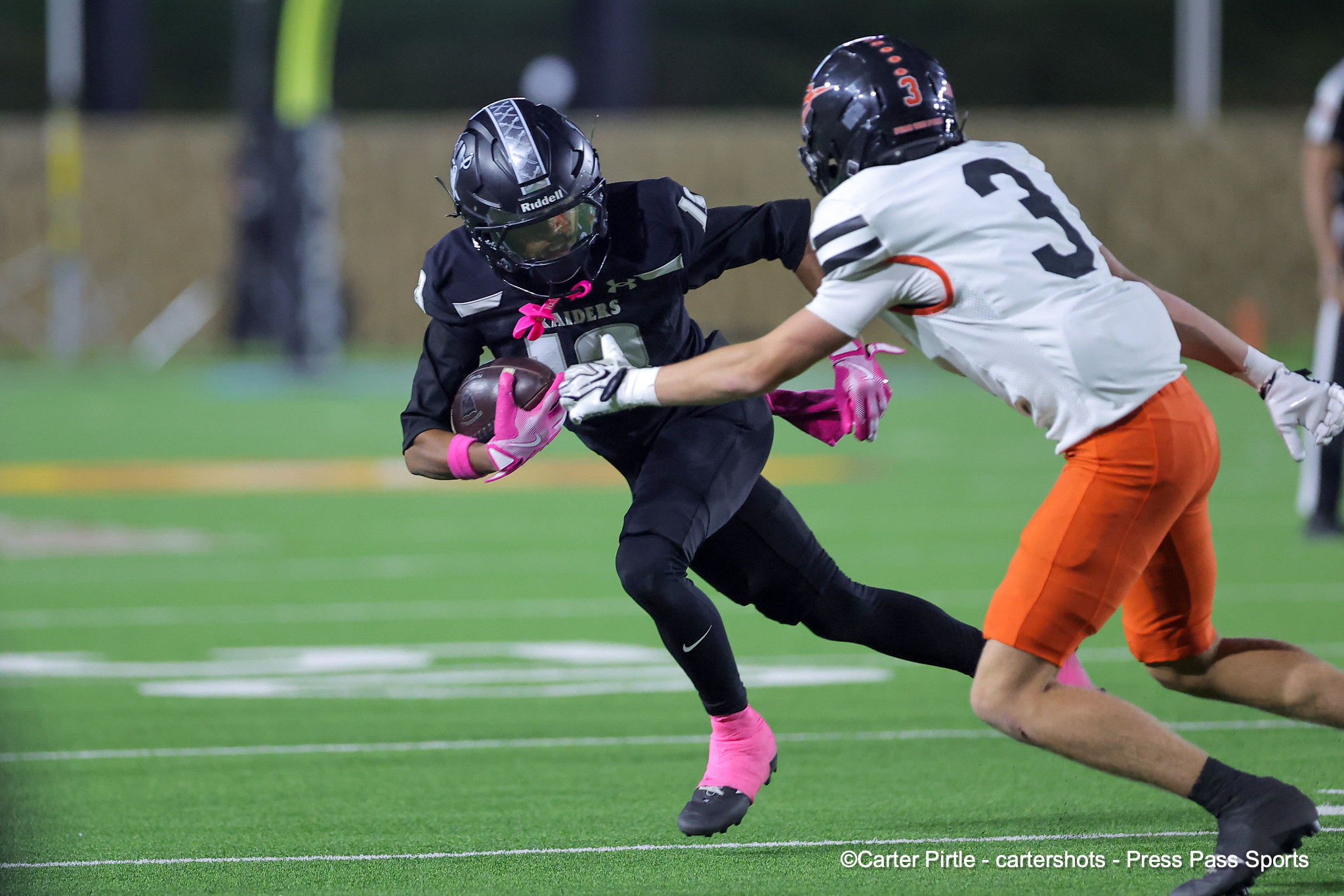 Two football players sprint on a green turf; a Black-uniformed player with a pink-gloved hand carries the ball while a white-uniformed opponent pursues him.