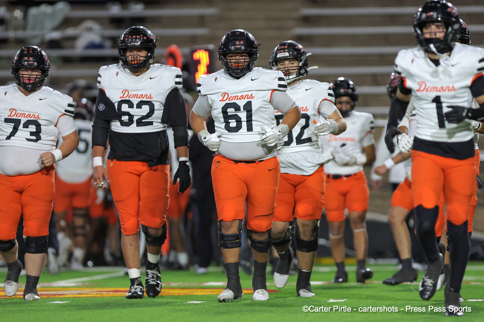 College football players in white Demons jerseys and orange pants walking on the field with helmets.