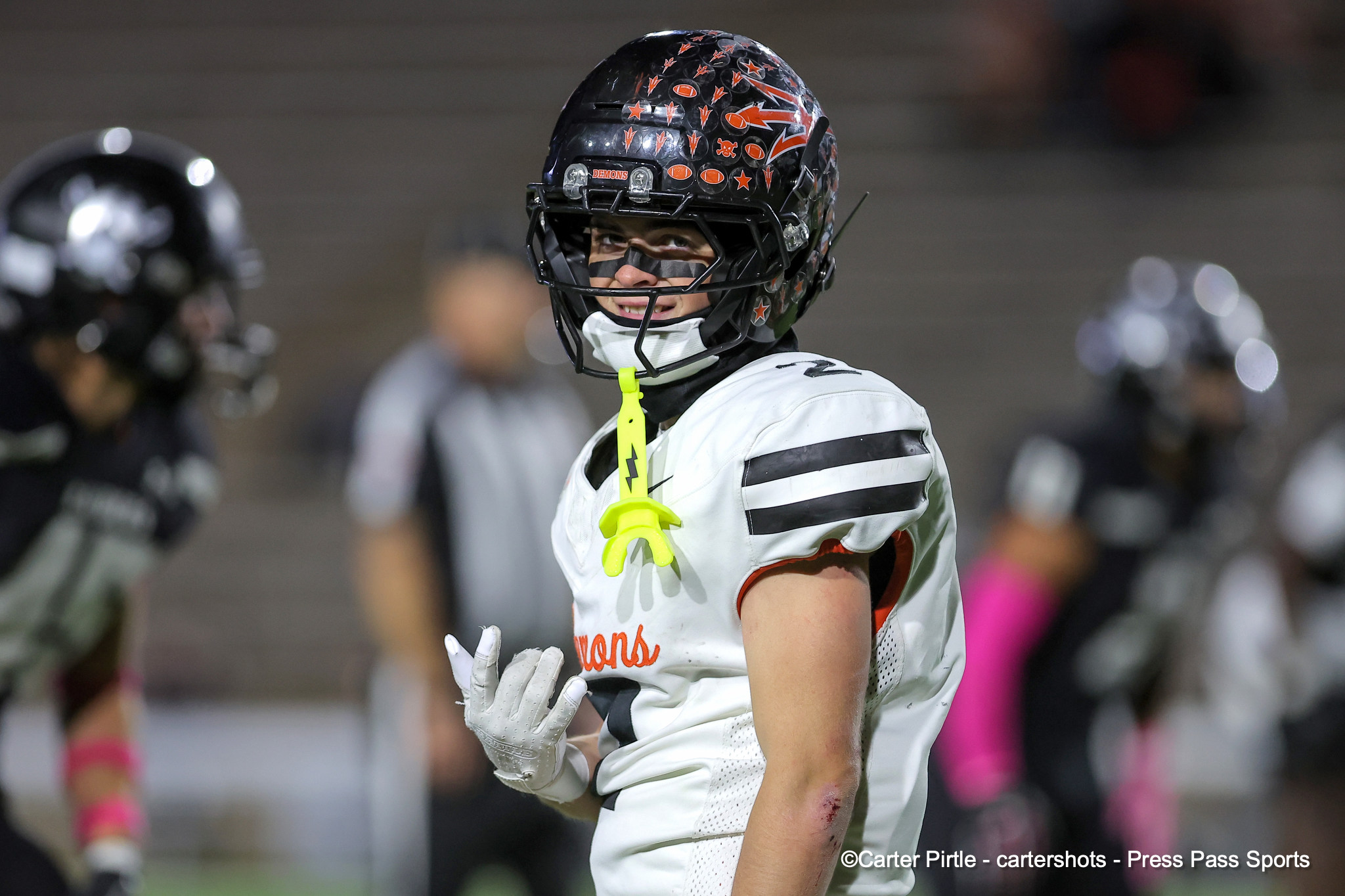 Lacrosse player in a white jersey with a decal-covered helmet and neon-yellow mouthguard at front.